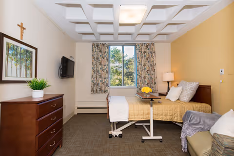 A furnished senior care bedroom with a bed and overbed table, dresser, wall-mounted TV, window with floral curtains, and seating.