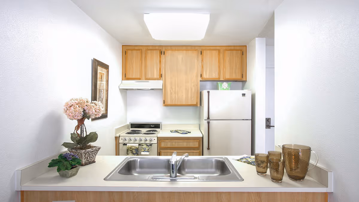 A compact kitchen with light wood cabinets, a white refrigerator, an electric stove with oven, and a double stainless steel sink in the foreground. The countertop has a decorative flower arrangement on the left and a set of amber-colored glass pitcher and glasses on the right. A framed picture hangs on the left wall.