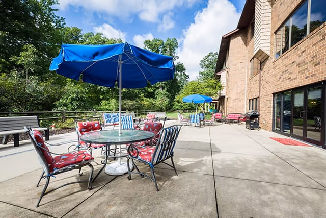 Outdoor patio area with round tables and chairs featuring red and blue cushions, blue umbrellas providing shade, a brick building on the right, and green trees in the background under a partly cloudy sky.
