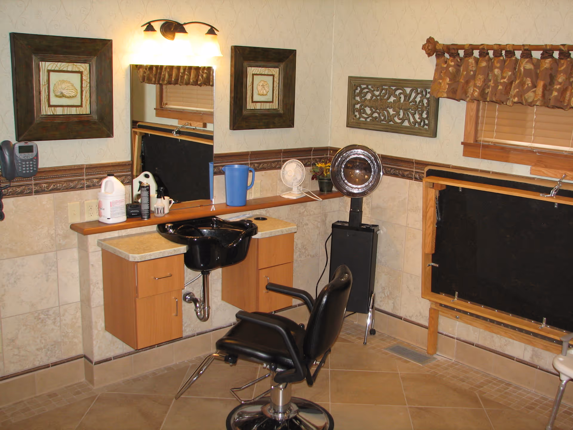 Interior view of a hair salon area with a black salon chair in front of a black sink mounted on a beige countertop with wooden cabinets. Above the sink is a large mirror with two framed pictures on either side and a light fixture above. There is a small fan and a potted plant on the countertop, and a window with a brown valance on the right side.