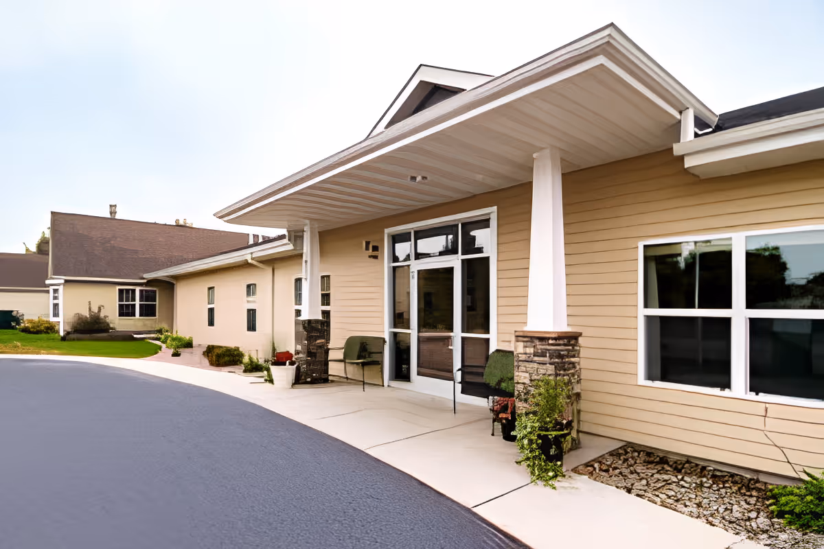 Exterior view of Edgewood Prairie Crossings Assisted Living facility showing the entrance with a covered porch supported by white columns with stone bases, beige siding, large windows, and a paved driveway.