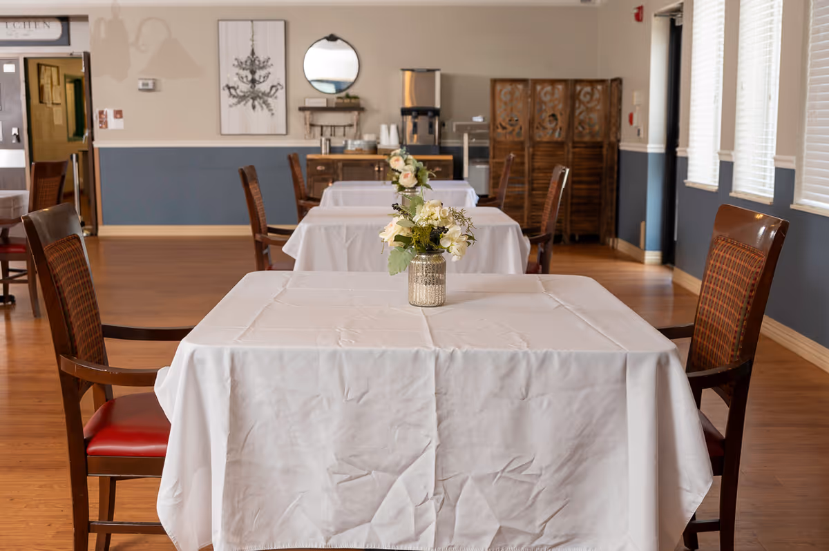 A dining room with tables covered in white tablecloths, each adorned with a small floral centerpiece. Wooden chairs with red cushions surround the tables. The room has wooden flooring, blue and beige walls, and windows with white blinds on the right side. In the background, there is a wooden cabinet, a coffee machine, and a decorative mirror on the wall.