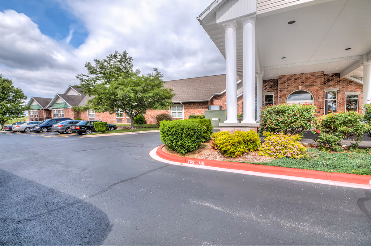 Brick senior living building entrance with white columns, landscaped shrubs and a parking lot.