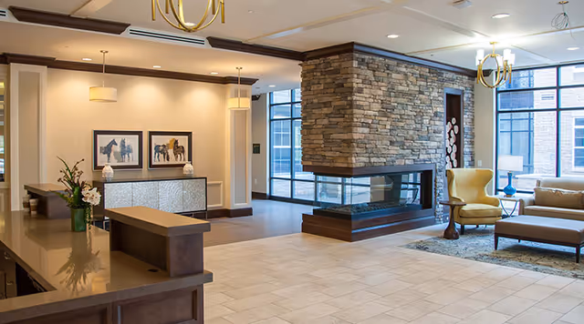 A spacious and well-lit interior common area featuring a stone fireplace in the center, modern chandeliers, large windows letting in natural light, a yellow armchair, a beige sofa, a wooden coffee table, and a reception desk with a flower vase and decorative items.