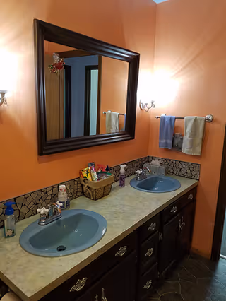 Bathroom with two blue sinks set in a long countertop with a mosaic tile backsplash. Above the sinks is a large rectangular mirror with a dark wooden frame. The walls are painted orange, and there are two wall-mounted light fixtures on either side of the mirror. A towel rack with two towels, one blue and one beige, is mounted on the wall to the right.