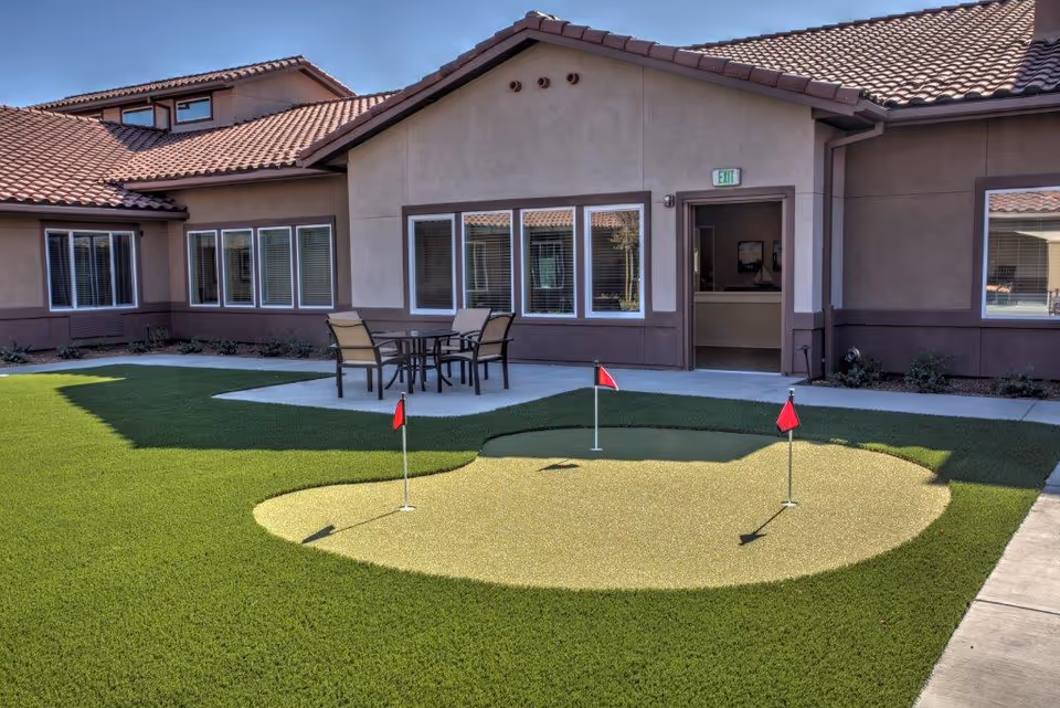 Outdoor courtyard area at Vineyard Place featuring a small putting green with three red flags, surrounded by artificial grass. There is a concrete patio with a table and four chairs near the building entrance, which has multiple windows and a door with an exit sign above it.