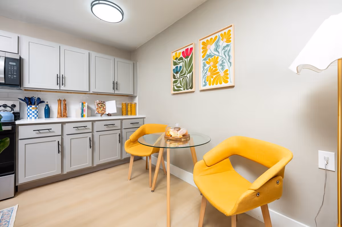 A modern kitchen and dining area featuring light gray cabinets with black handles, a glass-top round table with wooden legs, and two bright yellow upholstered chairs. On the countertop are kitchen utensils, cookbooks, and jars of pasta. Two colorful floral paintings hang on the wall above the dining table. A floor lamp is partially visible on the right side.