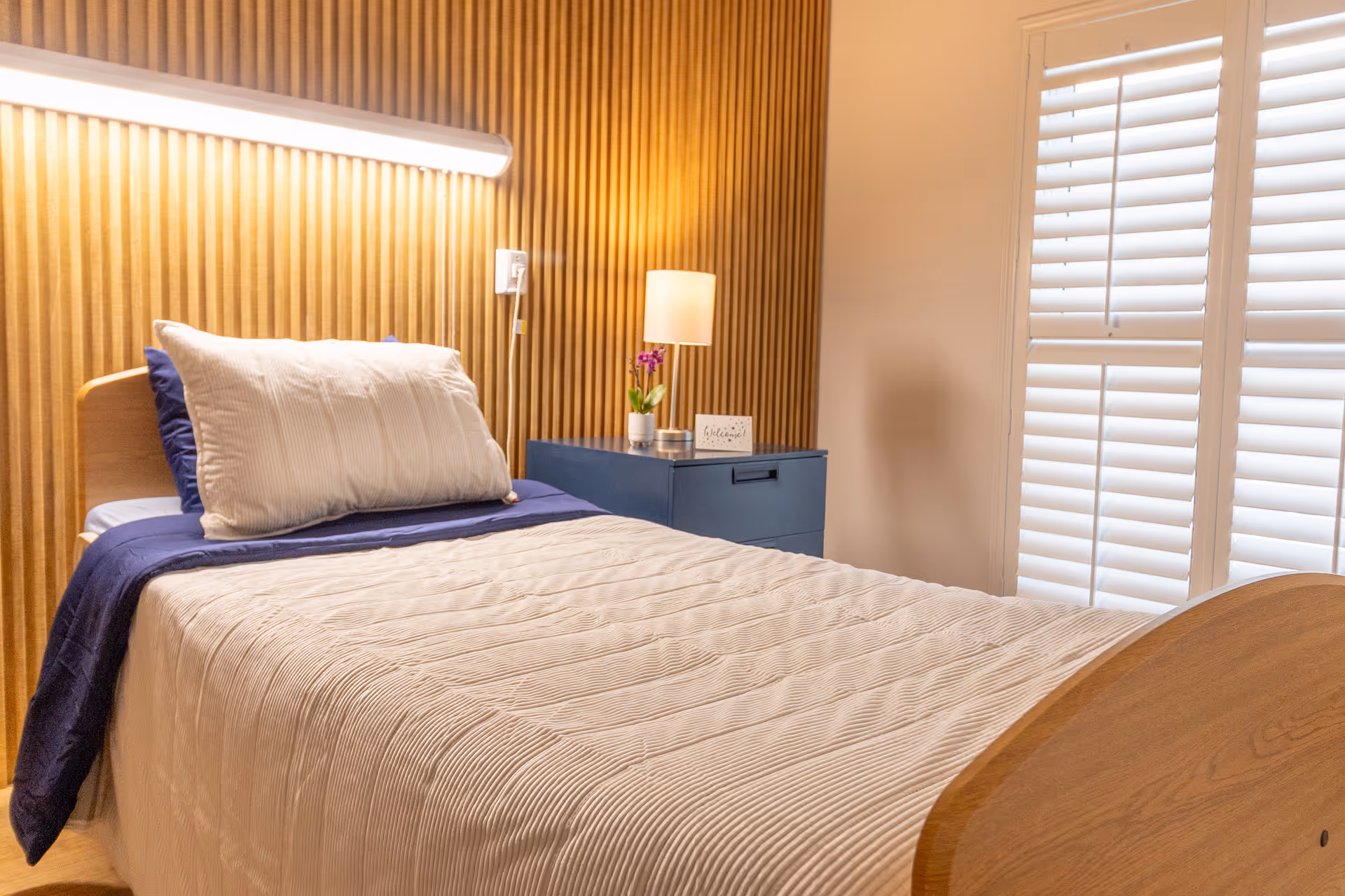 A neatly made single bed with a white pillow and beige quilt in a room with a wooden slat accent wall. Next to the bed is a blue nightstand with a small lamp, a potted plant, and a decorative sign. White plantation shutters cover the window, allowing soft natural light into the room.