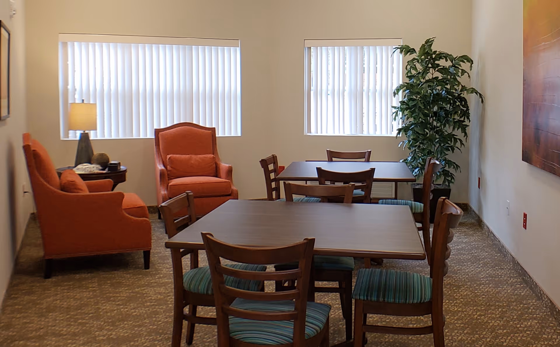 A cozy room with two orange armchairs and a small side table with a lamp on the left side. There are two rectangular tables with wooden chairs that have green striped cushions. Two windows with vertical blinds let in natural light. A tall green plant is placed in the corner on the right side, and a large abstract painting hangs on the right wall.