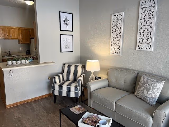 A cozy living room area with a gray sofa adorned with a patterned pillow, a striped armchair, a small black coffee table with a tray and a cup, and a side table with a lamp. The wall behind the sofa features two decorative white panels and two framed black and white floral prints. The room has wooden flooring and an open view into a kitchen area with wooden cabinets.