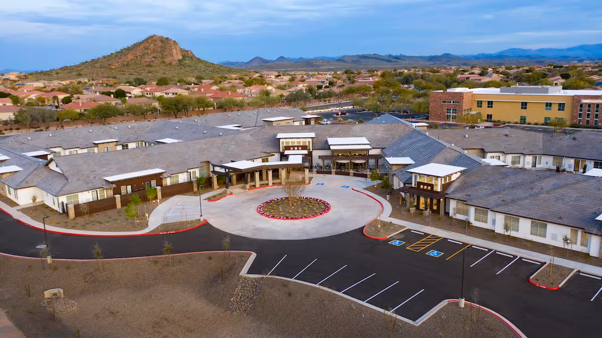 Aerial view of Spring Gardens Senior Living Peoria facility showing a large single-story building with a circular driveway and landscaped center island. Surrounding the building are parking spaces including handicapped spots. In the background, there are residential houses and a mountain under a partly cloudy sky.