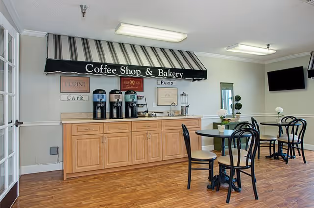 Interior view of a coffee shop and bakery area in a senior living facility with wooden cabinets, three coffee dispensers, a water dispenser, and small round tables with black chairs. The wall has a black awning sign that reads 'Coffee Shop & Bakery' along with decorative signs and a mounted flat-screen TV.