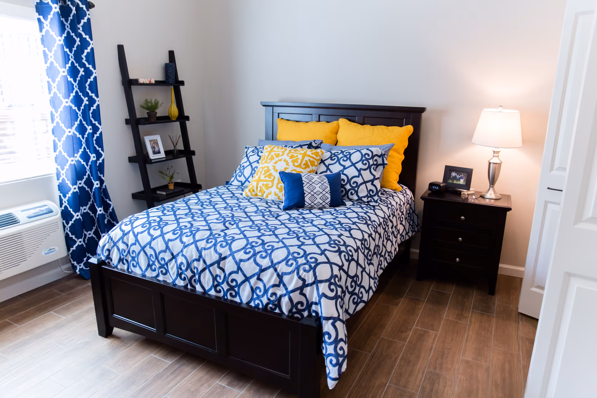 A neatly made bed with a dark wooden frame and blue and white patterned bedding, accented with yellow and blue pillows. To the right of the bed is a dark wooden nightstand with a lamp, a framed photo, and a clock. To the left is a window with blue and white patterned curtains and a black ladder-style shelf holding decorative items. The room has wooden flooring and light-colored walls.
