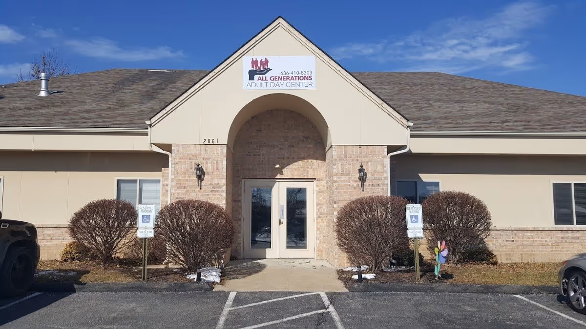 Front exterior view of a single-story building with a peaked roof and a central entrance featuring double glass doors. The building has beige siding with brick accents around the entrance. There are two handicap parking signs in front of the building, along with trimmed bushes on either side of the entrance. A sign above the entrance reads 'All Generations Adult Day Center' with a phone number.