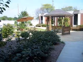 Outdoor courtyard with a gazebo, benches, umbrella, and planted landscaping at a senior facility.