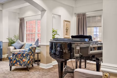 Communal living room with a black grand piano, patterned armchairs, potted plants and large windows.