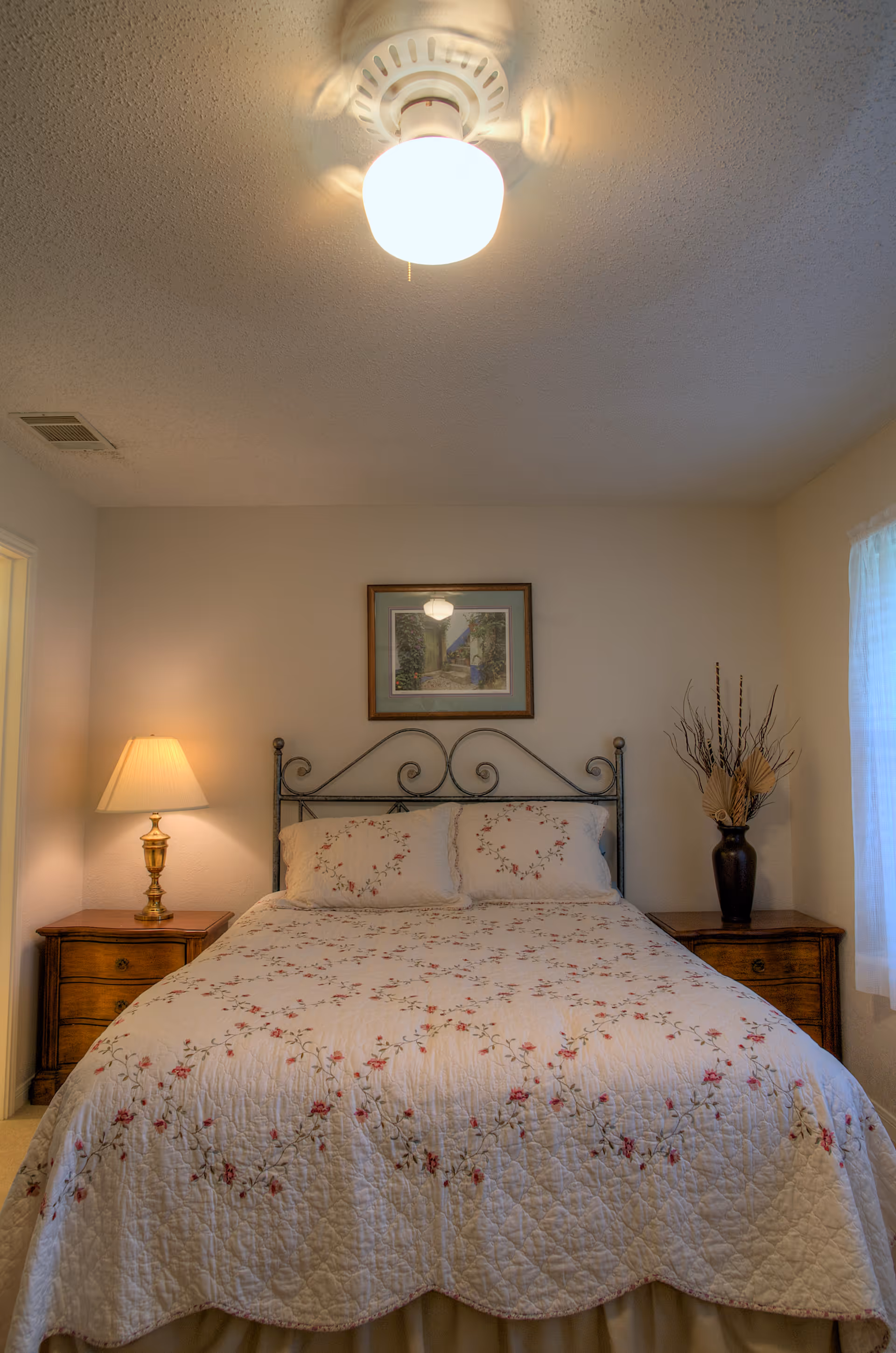 A neatly made bed with a floral quilt and pillows between two wooden nightstands, a lamp on the left, a vase on the right, a framed picture above, and a ceiling light.