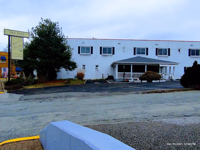 Exterior view of a two-story white building with several windows and a covered porch entrance. There is a sign on the left side that reads 'Gracious Living Personal Care Home' along with a phone number. The surrounding area includes a paved parking lot, some bushes, and trees under a cloudy sky.