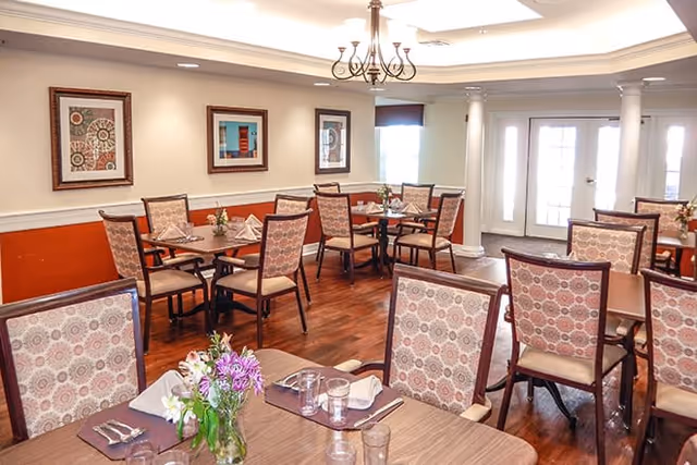 A well-lit dining room with multiple wooden tables and chairs featuring patterned upholstery. Each table is set with placemats, napkins, glasses, and small floral centerpieces. The room has hardwood flooring, framed artwork on the walls, and a chandelier hanging from the ceiling. Large windows and glass doors allow natural light to enter the space.