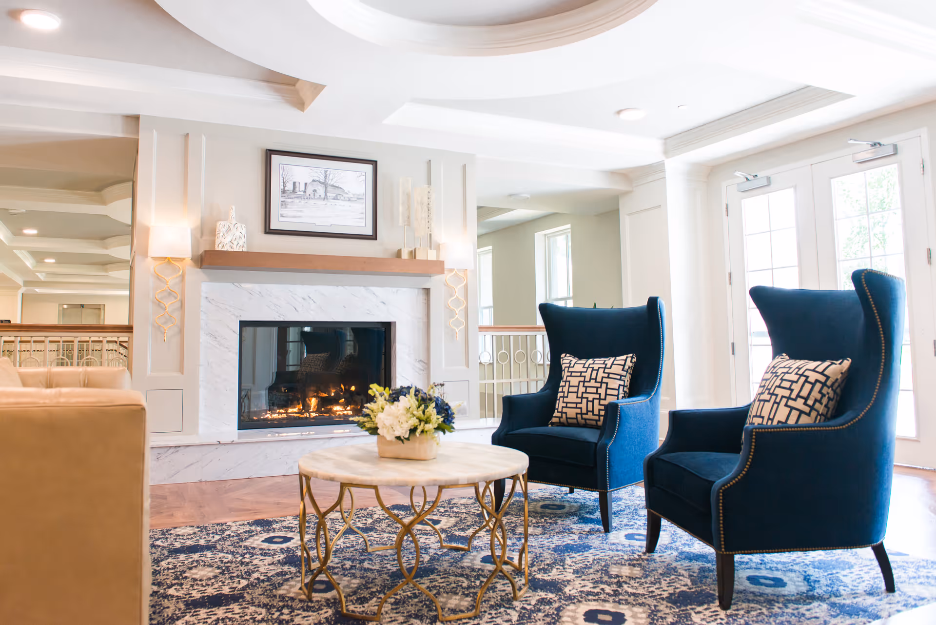 A bright and elegant living room area featuring a lit fireplace with a white marble surround, two dark blue armchairs with patterned cushions, a round marble-top coffee table with a floral arrangement, and a beige sofa partially visible on the left. The room has large windows and glass doors allowing natural light to fill the space.