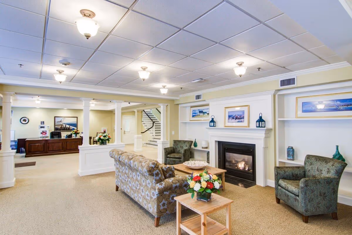 A bright and spacious living room area in Crescent Point at Niantic featuring a patterned sofa, two upholstered armchairs, a wooden coffee table with a decorative bowl, and a small side table with a flower arrangement. The room has a fireplace with framed pictures and decorative lanterns on the mantel, built-in shelves with vases, and a view into a reception desk area with wooden furniture and floral decorations. The ceiling has multiple light fixtures, and there is a staircase visible in the background.