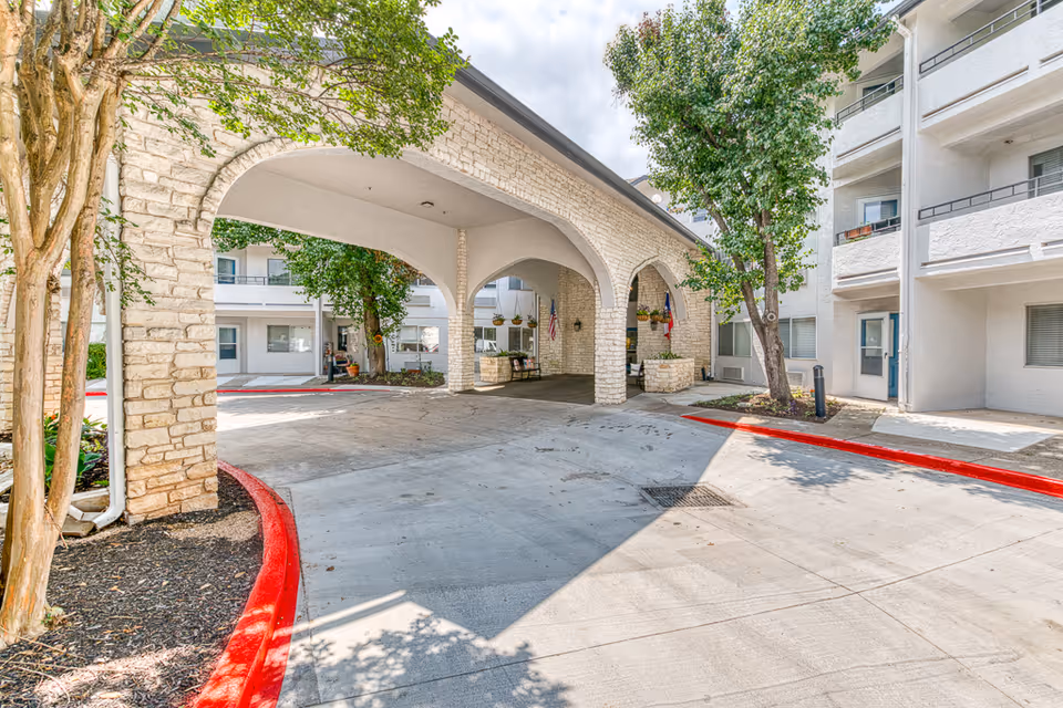 Entrance area of a senior living facility with a covered driveway supported by stone arches. The building is white with multiple balconies and windows. There are trees and landscaping around the driveway, and red-painted curbs line the edges of the pavement.