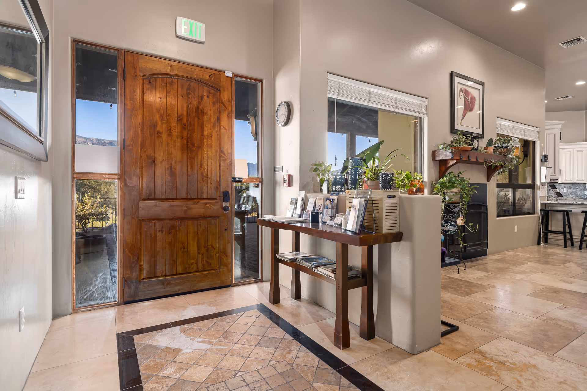 Interior view of an assisted living facility entrance area with a large wooden front door, a table displaying brochures and plants, a window with blinds, and a fireplace with a mantel decorated with plants and a framed picture.