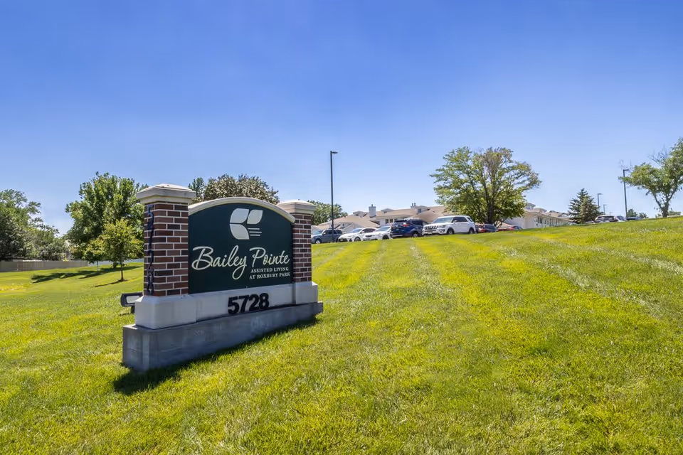 Outdoor view of Bailey Pointe at Roxbury Park assisted living facility sign on a grassy hill with parked cars and trees in the background under a clear blue sky.
