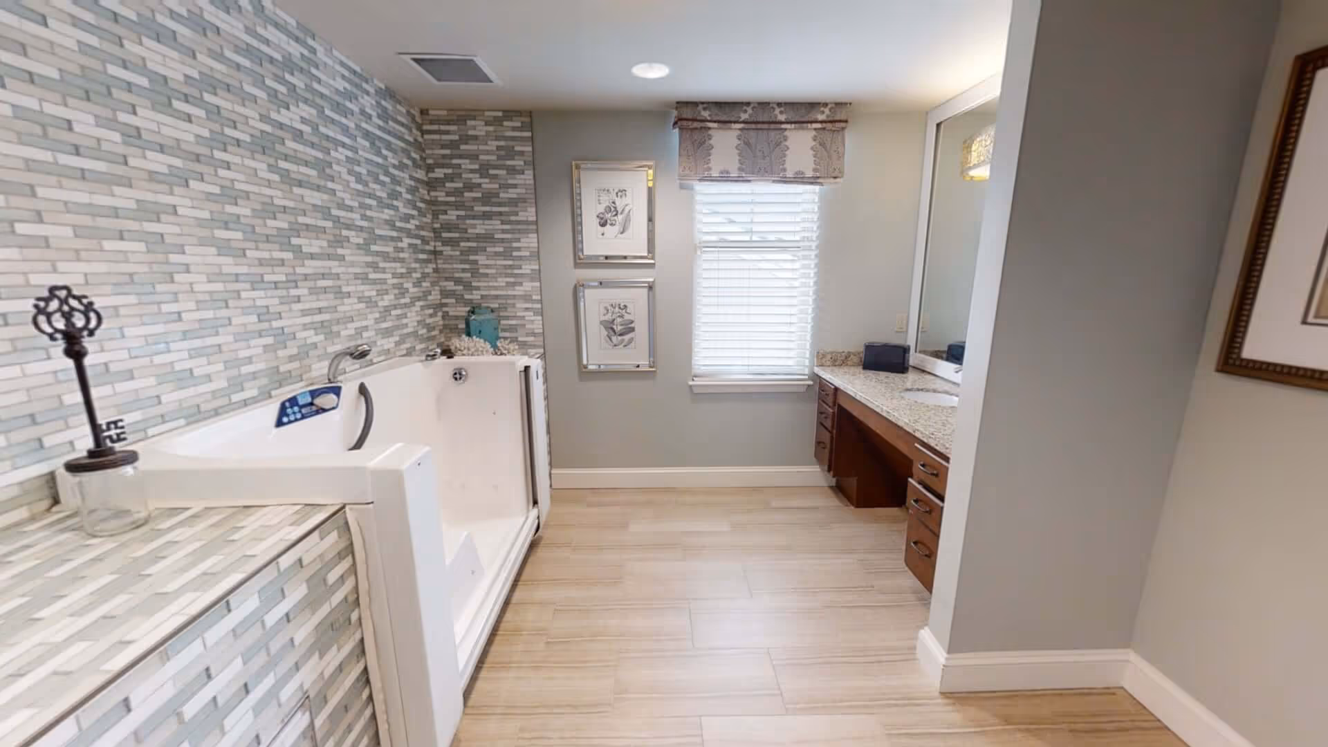A modern bathroom featuring a walk-in bathtub with a door, surrounded by a mosaic tile wall. The room has a large window with blinds and a decorative valance, two framed botanical prints on the wall, a granite countertop with a sink, and wooden cabinetry underneath. The floor is covered with light-colored tiles.