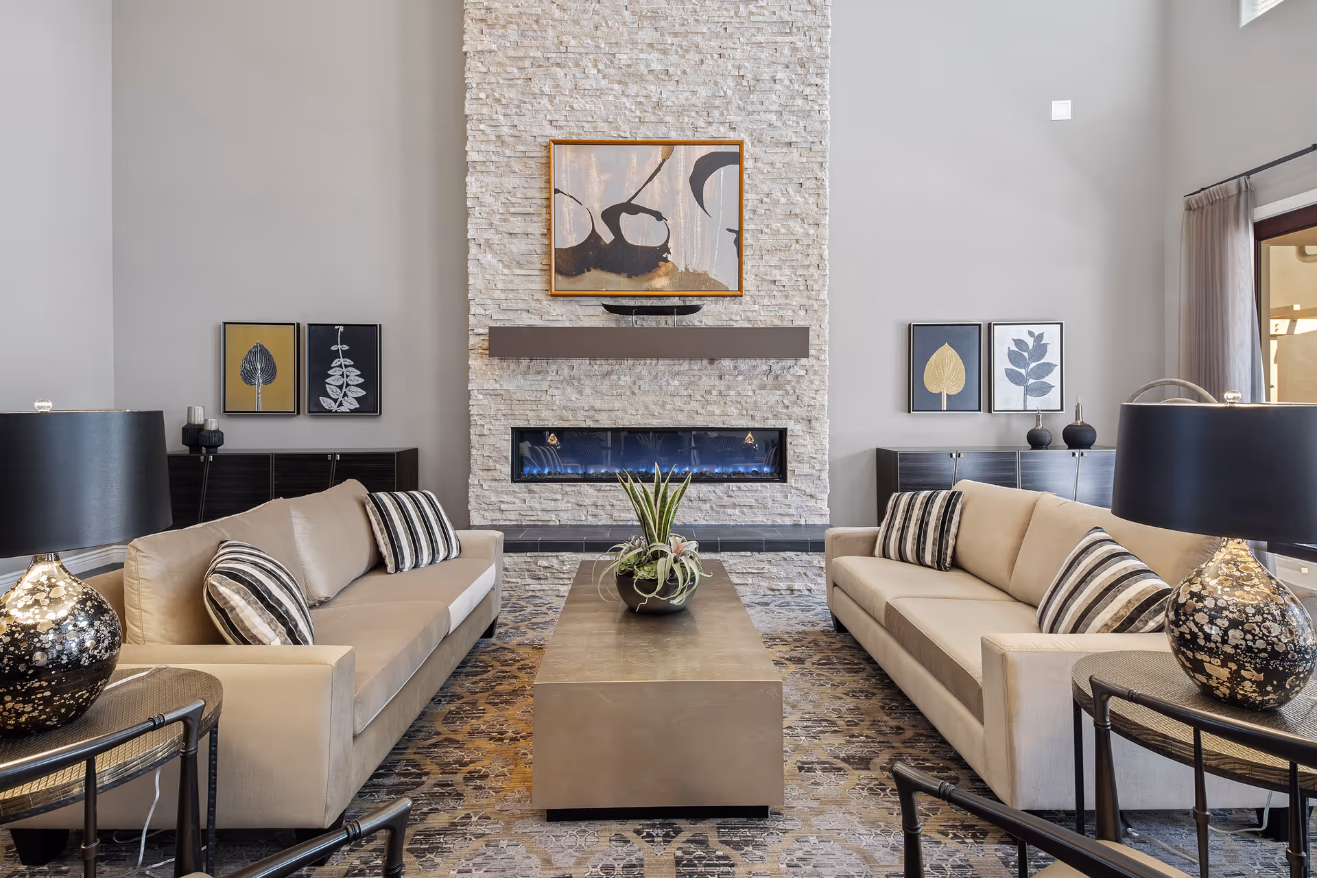 A modern living room with two beige sofas facing each other, each adorned with striped pillows. Between the sofas is a rectangular coffee table with a plant centerpiece. The room features a tall stone fireplace with a contemporary abstract painting above it. On either side of the fireplace are dark wood cabinets with framed botanical artwork above them. Two decorative table lamps with black and gold bases are placed on side tables next to the sofas. The room has high ceilings and large windows with sheer curtains allowing natural light to enter.