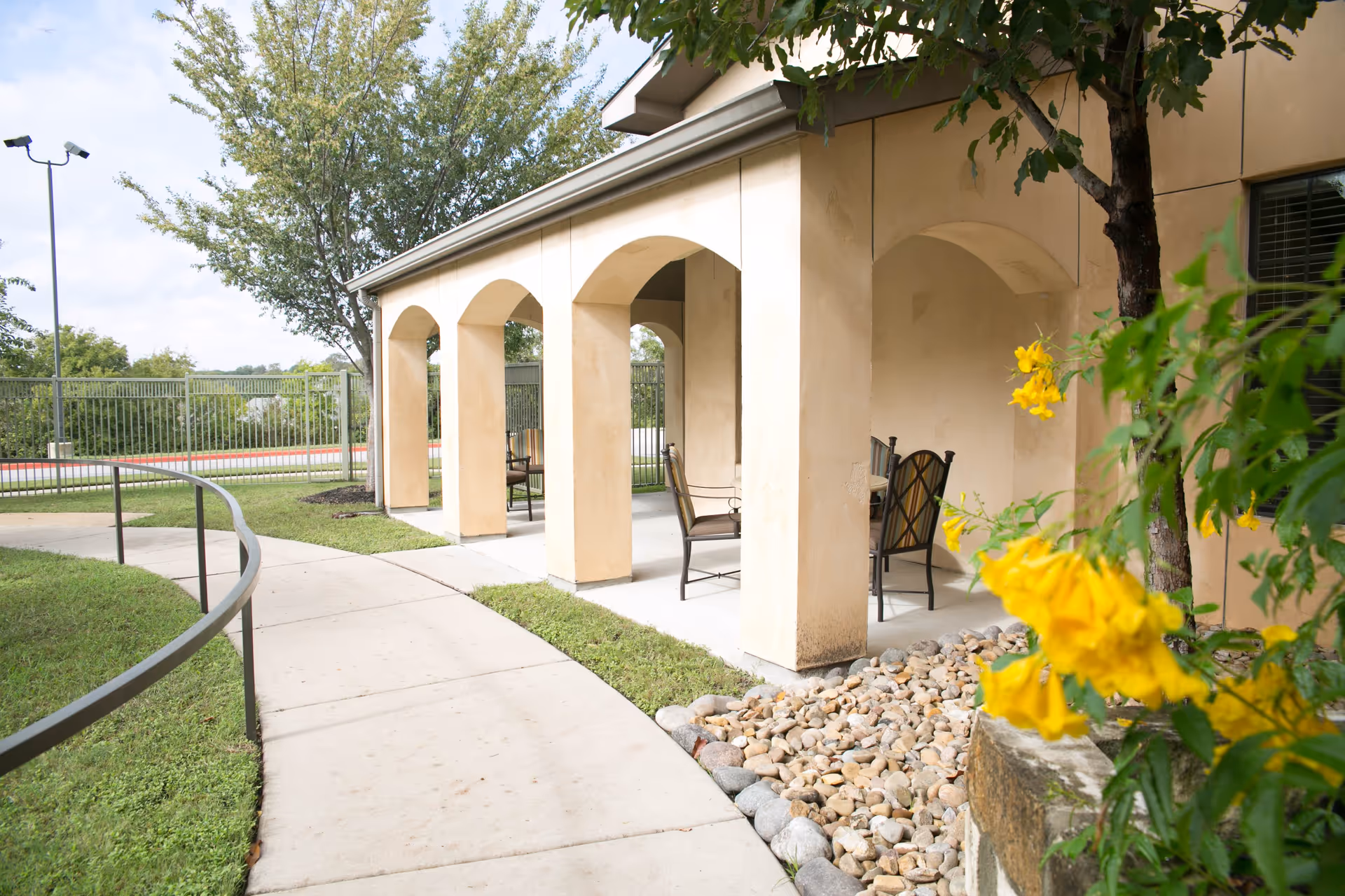 Curved concrete walkway leading to a covered arched patio with chairs, rock landscaping, trees, and yellow flowers.