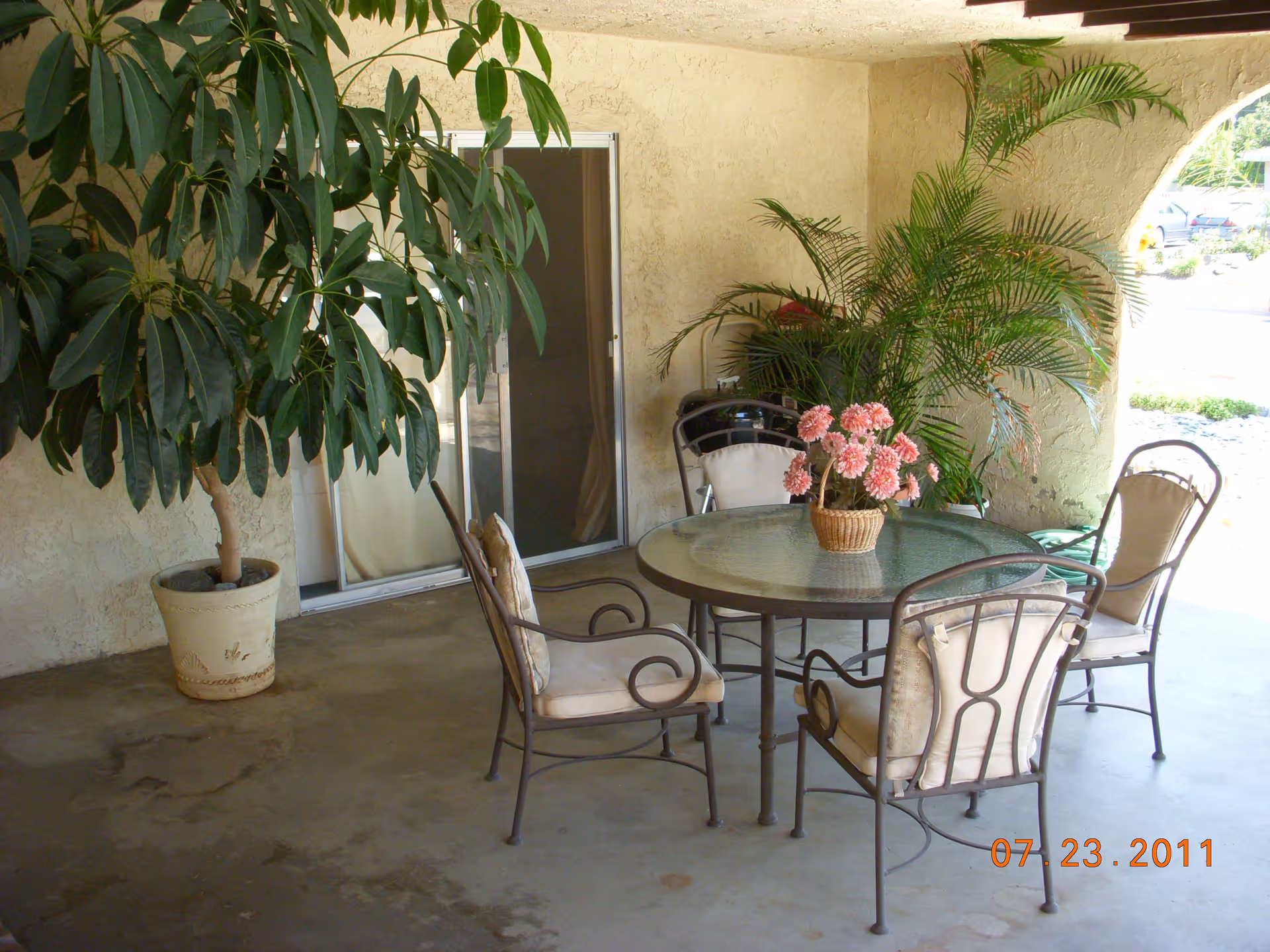Covered outdoor patio area with a round glass table surrounded by four cushioned metal chairs. There is a potted plant with pink flowers on the table and large green potted plants near the wall. A sliding glass door is visible in the background.