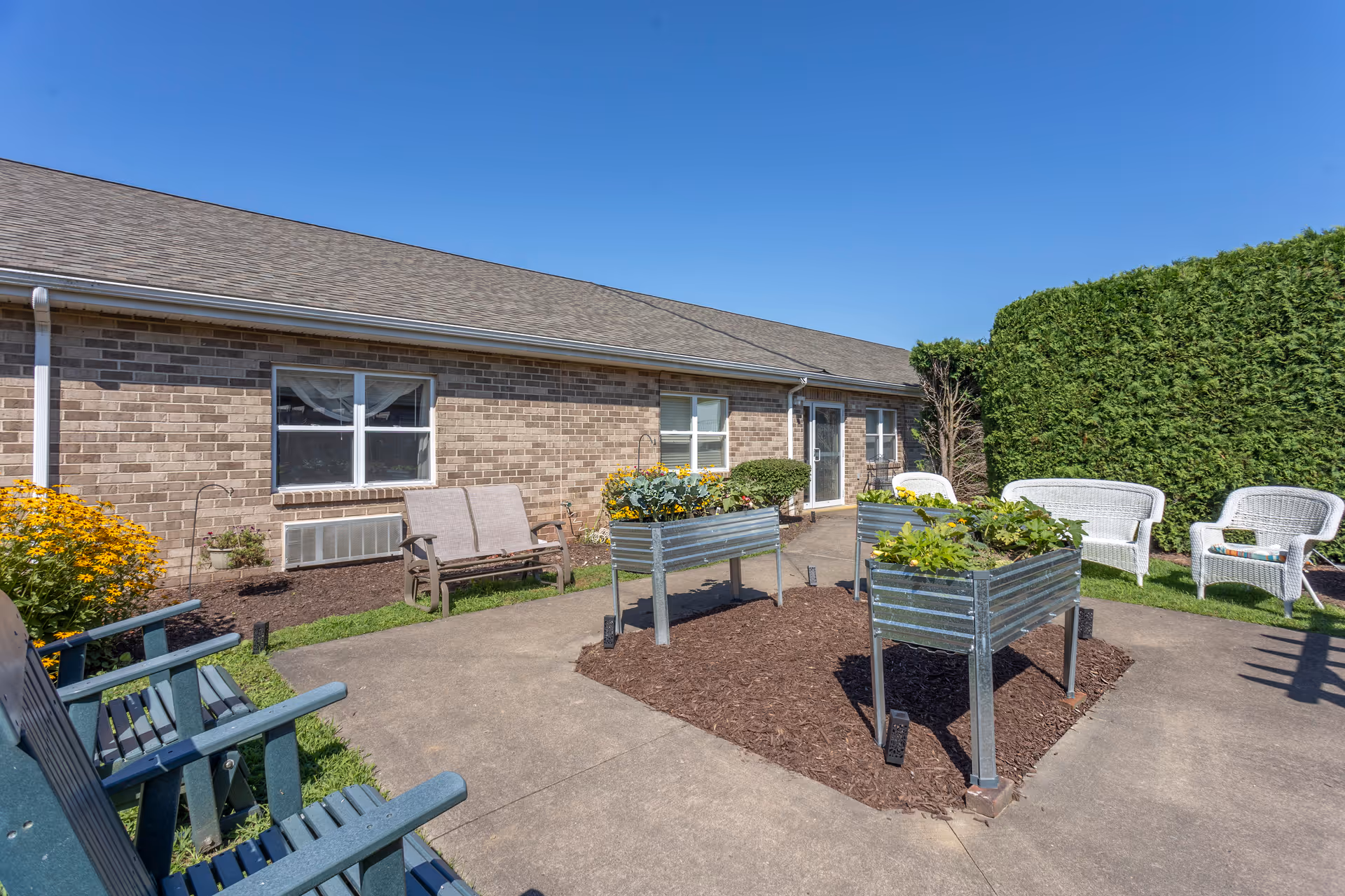 Outdoor patio area at Brookdale Bloomsburg featuring raised garden beds with plants, surrounded by various seating options including blue wooden chairs, a bench, and white wicker chairs. The patio is adjacent to a brick building under a clear blue sky.