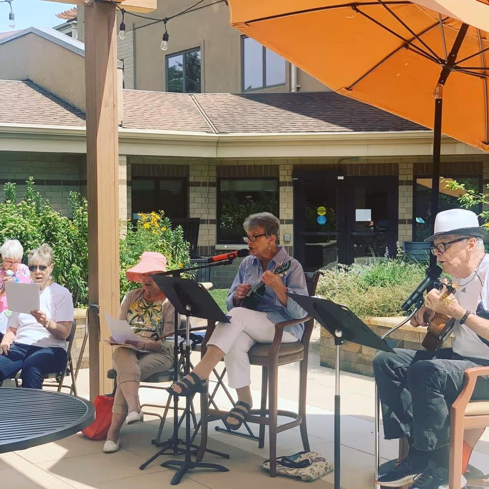 A group of elderly people sitting outdoors under a large orange umbrella at Harwood Place Senior Living. Two individuals are playing string instruments and singing into microphones, while others are seated nearby holding papers, possibly song lyrics. The setting is a patio area with greenery and a building in the background.