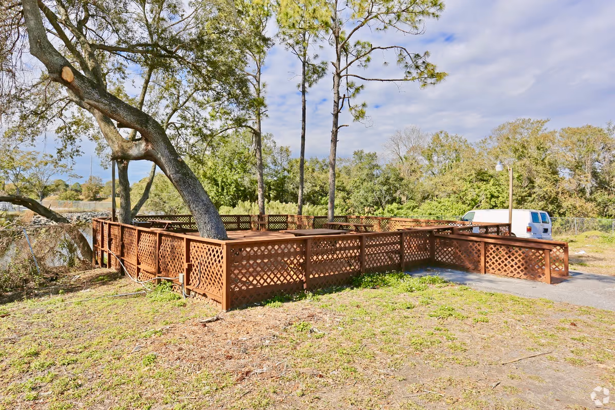Outdoor area with a wooden lattice fence surrounding a tree and a paved path leading to the fenced area. There is greenery and trees in the background, along with a white van parked near the fence.