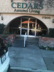 Entrance to Cedars Assisted Living facility with a glass door under an archway, flanked by brick planters and a ramp with handrails leading up to the door. The building number 17300 is visible on the right side of the entrance.