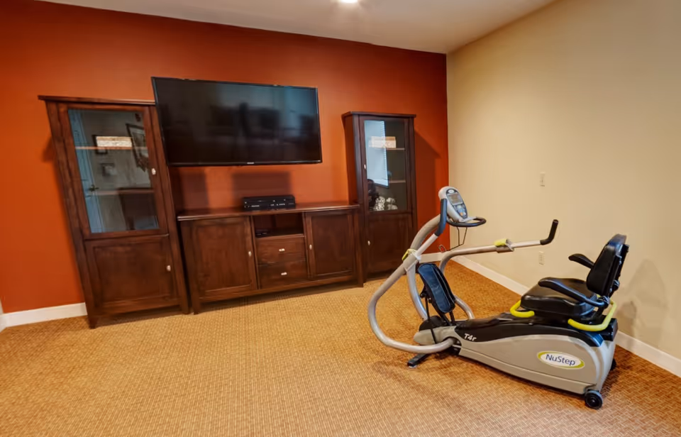 A room with a NuStep exercise machine on a carpeted floor, a large flat-screen TV mounted on a burnt orange wall, and a wooden entertainment center with glass cabinets on either side.