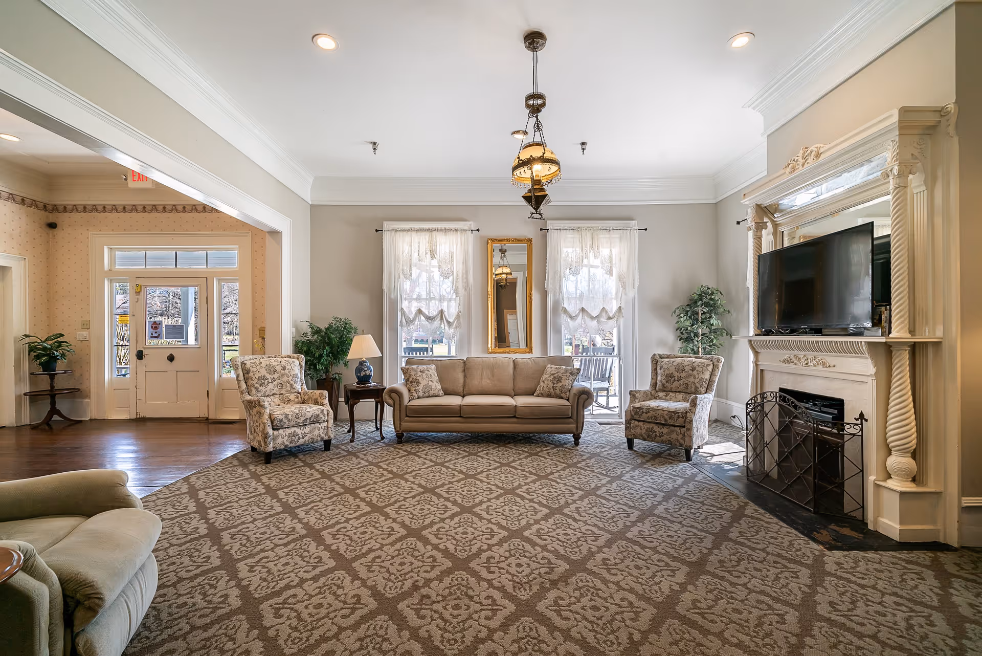 Bright communal living room with a sofa, patterned armchairs, a fireplace with TV, and large windows.