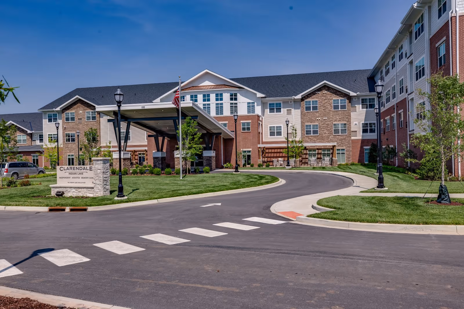 Exterior view of Clarendale at Indian Lake, a senior living facility with a large multi-story building featuring brick and siding, a covered entrance with an American flag, a curved driveway, street lamps, and well-maintained landscaping under a clear blue sky.