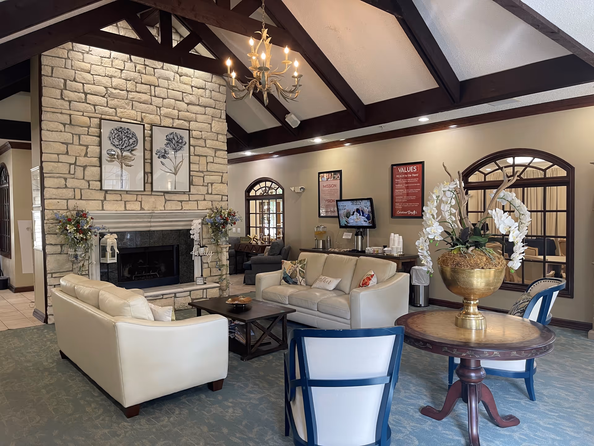 A cozy living room area in a senior living facility with two cream-colored sofas facing a stone fireplace adorned with floral artwork and decorative flower arrangements. The room features a high ceiling with exposed dark wooden beams and a chandelier. There is a round wooden table with a large gold vase holding white flowers and decorative branches, and two blue and white chairs nearby. In the background, a TV is mounted on the wall above a table with refreshments and informational posters.