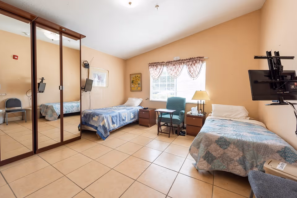 A bright assisted-living bedroom with two twin beds, mirrored closet doors, a window with an armchair and bedside tables, tiled floor and wall-mounted TVs.
