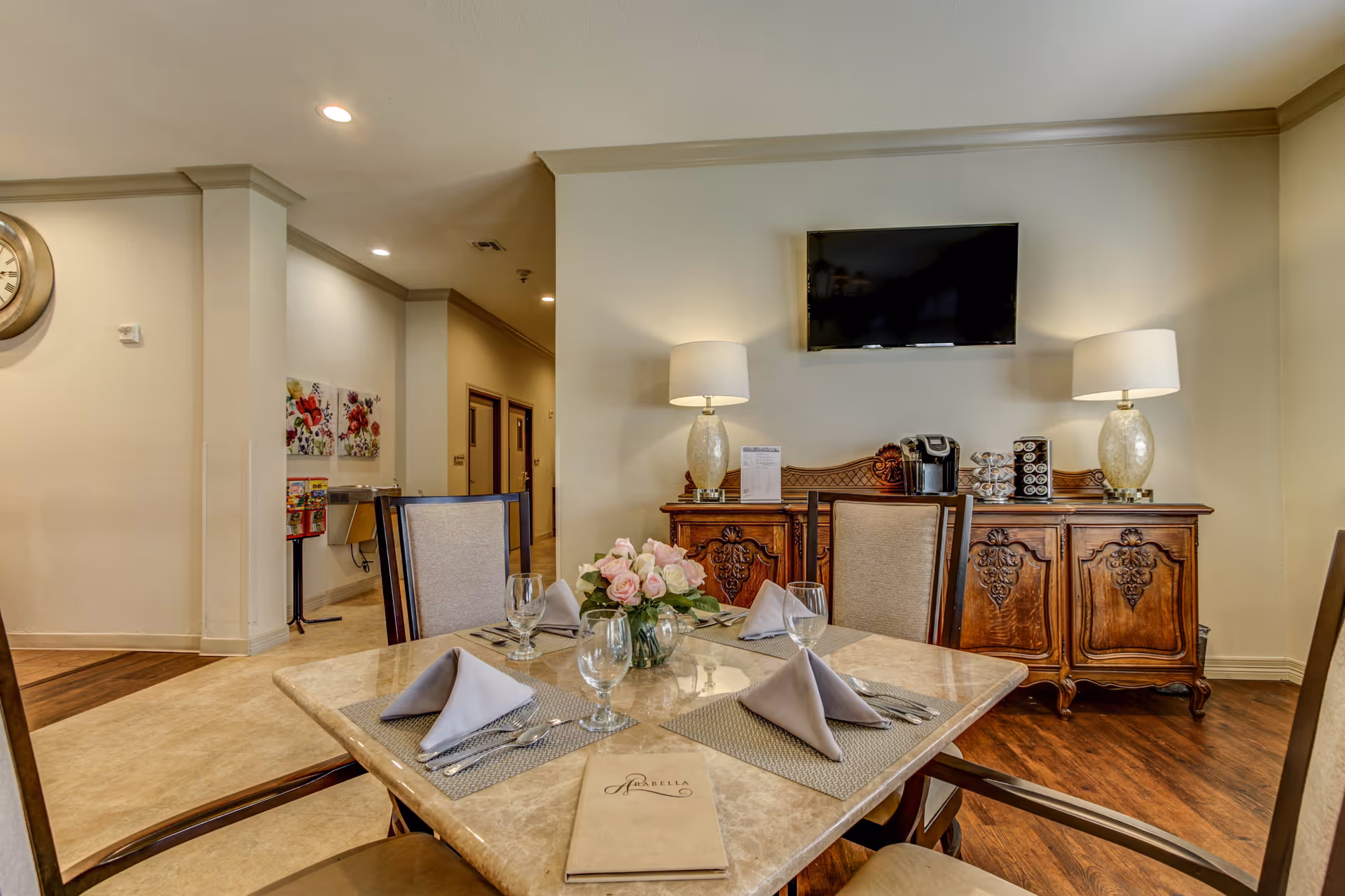A dining area in a senior living facility with a square marble table set for four, including folded napkins, glasses, and silverware. A vase with pink roses is in the center of the table. Behind the table is a wooden sideboard with ornate carvings, two table lamps, a coffee maker, and a mounted flat-screen TV above it. The room has warm lighting and a hallway is visible in the background.