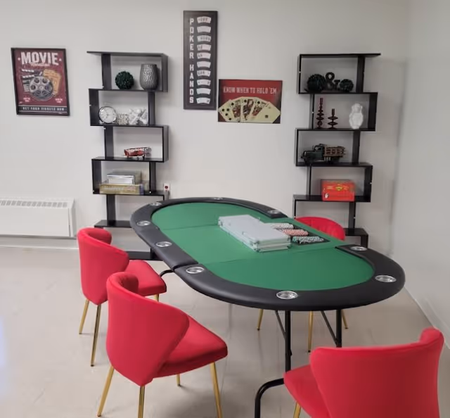 A poker table with green felt surrounded by pink chairs in a small game room with shelving and poker-themed wall signs.