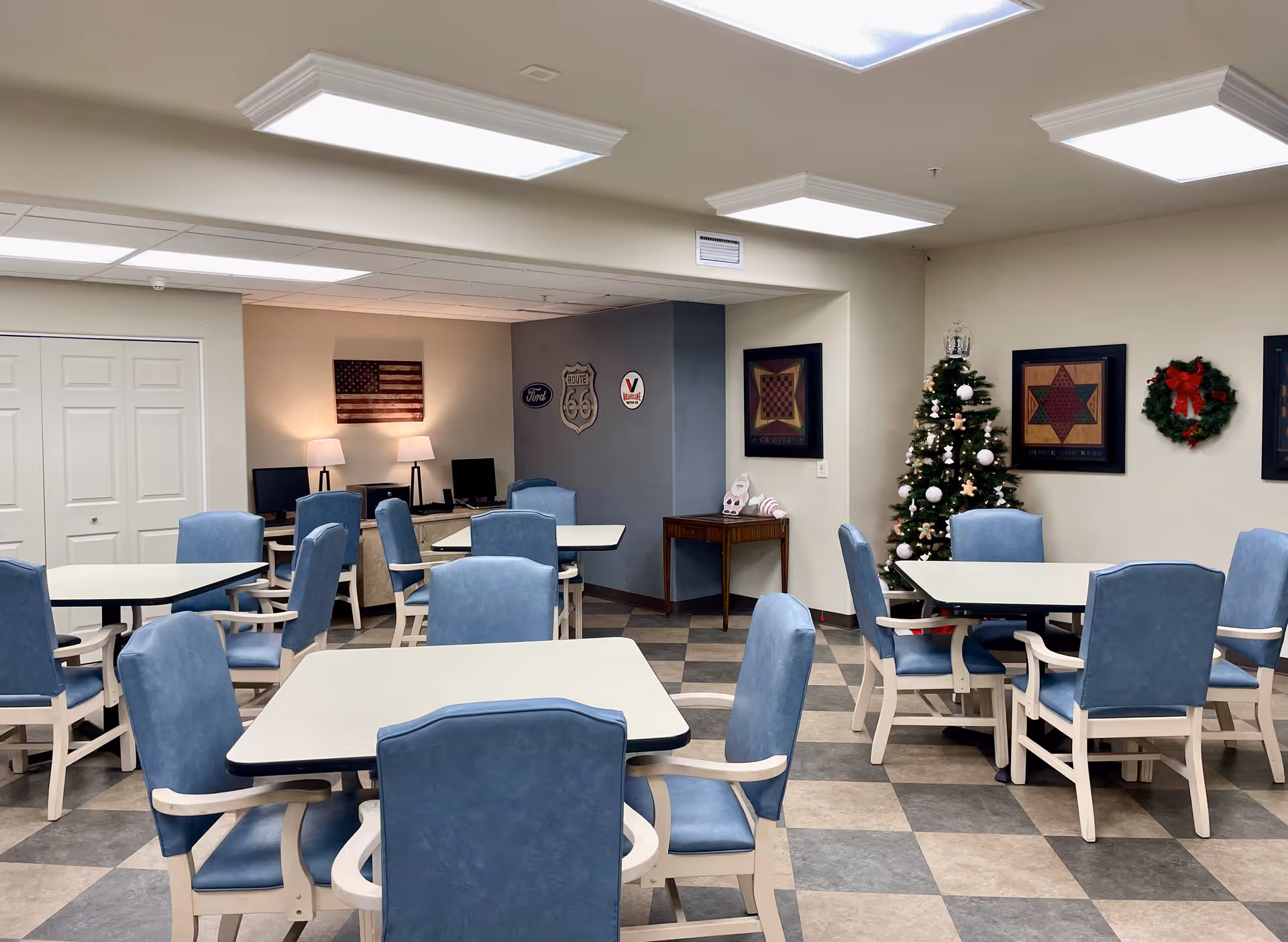 A well-lit common room with several tables and blue cushioned chairs arranged neatly. The room features a decorated Christmas tree, framed artwork, a wreath on the wall, and a small table with holiday decorations. In the background, there are computer stations with two monitors and an American flag wall decoration.