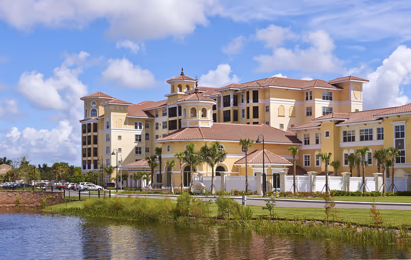 Large Mediterranean-style multi-story senior living building with red tile roofs, palm trees, and a reflecting pond in the foreground.