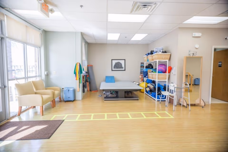 Bright therapy/exercise room with two armchairs by large windows, a treatment table, shelves of exercise equipment, and agility-ladder markings on the floor.