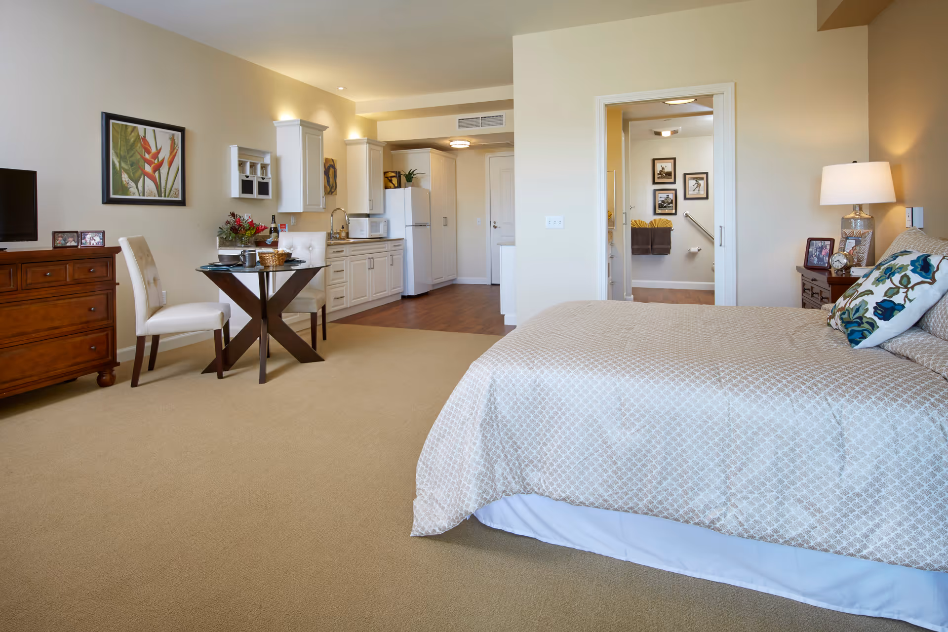 A spacious senior living facility room at 'Ilima at Leihano featuring a bed with patterned bedding and decorative pillows, a wooden nightstand with a lamp and framed photos, a small dining table with two chairs, a kitchenette with white cabinets, a refrigerator, and a microwave. The room has beige walls and carpet, with a doorway leading to a bathroom visible in the background.