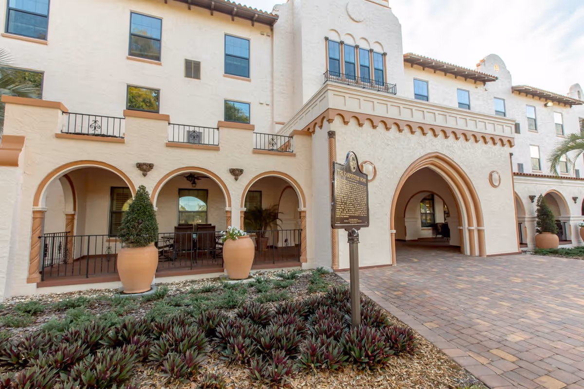 Exterior view of a large, light-colored building with arched doorways and windows, decorative columns, and a paved walkway. There are large potted plants and a historical marker sign in front of the building.