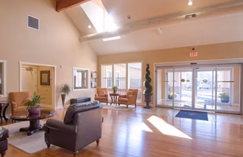 A spacious and well-lit common area with wooden floors, leather armchairs, a round table with a potted plant, large windows letting in natural light, and glass sliding doors leading outside. The ceiling has exposed wooden beams and skylights.