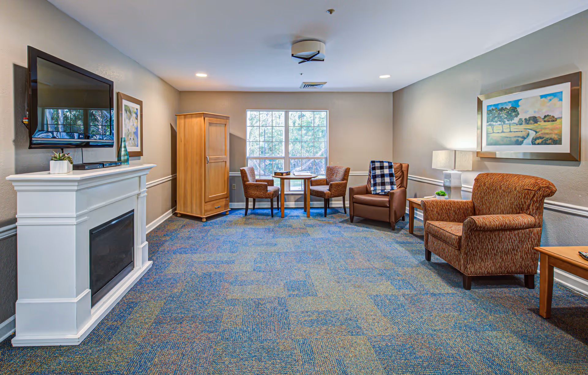 A cozy living room with a blue patterned carpet, a white electric fireplace with a flat-screen TV mounted above it, two framed paintings on the walls, a wooden armoire, a small round table with two chairs near a large window, and two upholstered armchairs with a side table and lamp.